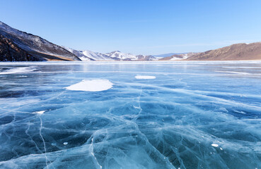 Beautiful winter landscape of frozen Baikal Lake on cold sunny day. Shallow bay is covered with blue ice with cracks against backdrop of  coastal mountains. Winter nature background. Copy space, blank