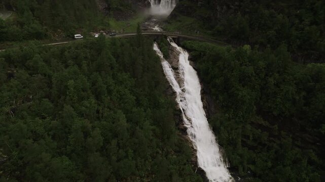 Aerial view of cascading Skjervefossen waterfall over lush green cliffs with a road and vehicles nearby, Granvin, Vestland, Norway.