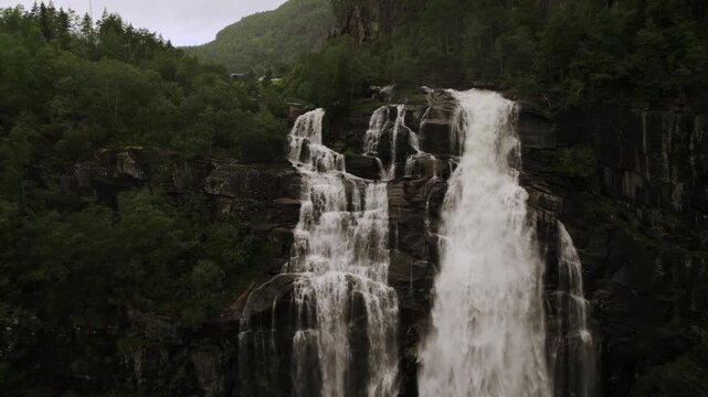 Aerial view of a cascading waterfall flowing down a rocky cliff face surrounded by lush green trees, creating a stark contrast, Granvin, Vestland, Norway.