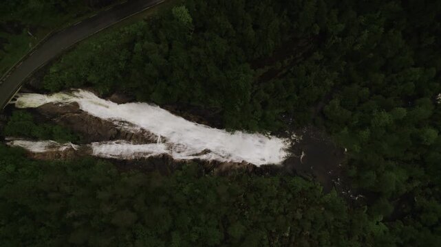 Aerial view of a cascading waterfall Skjervefossen cutting through a dense green forest, adjacent to a winding road, creating a stark contrast, Granvin, Vestland, Norway.