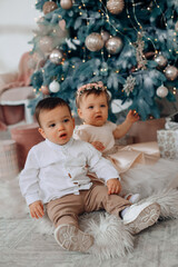 Fototapeta premium Children sitting on a soft rug next to a decorated Christmas tree in a cozy indoor space filled with holiday decorations and gifts