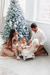 Family of four enjoys a cozy holiday moment by the Christmas tree with their toddlers playing with...