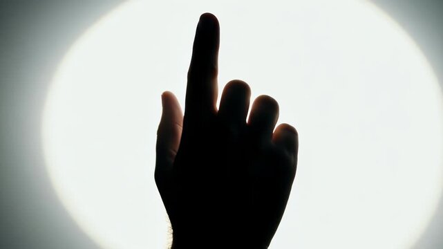 A silhouetted hand points toward a bright circular light, shifting slightly across frames as fingers adjust position against a clean illuminated background in a controlled studio setting.