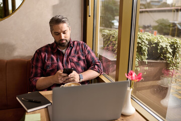 Mature man in stylish attire enjoys coffee while using smartphone in cozy cafe setting