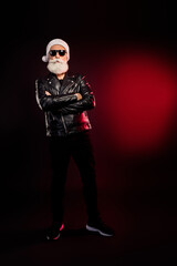 Cool Santa rocker in leather jacket with white beard and hat stands with arms crossed under red lights for holiday season photo shoot