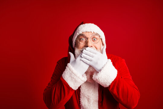 Santa in red suit with white beard and glasses covering mouth with gloved hands smiling against a bright red background - Powered by Adobe
