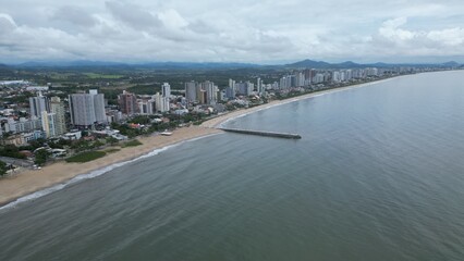 Aerial shot of a long pier jutting into a calm, gray sea under a cloudy sky. Unidentifiable people stand at the end, with distant islands on the horizon. Peaceful, minimalist coastal scene.