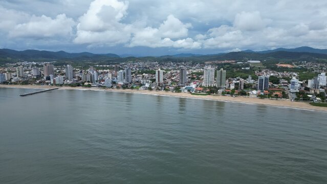 Aerial view of a coastal city with a long stone pier extending into the sea, sandy beach, and modern skyline against green hills under a cloudy sky. - Powered by Adobe