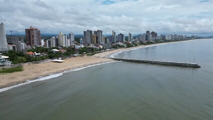 Aerial shot of a long pier jutting into a calm, gray sea under a cloudy sky. Unidentifiable people stand at the end, with distant islands on the horizon. Peaceful, minimalist coastal scene.
