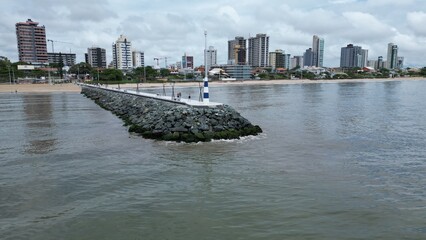 Aerial view of a coastal city with a long stone pier extending into the sea, sandy beach, and modern skyline against green hills under a cloudy sky.