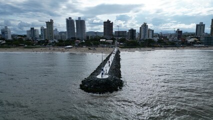 Aerial view of a coastal city with a long stone pier extending into the sea, sandy beach, and modern skyline against green hills under a cloudy sky.
