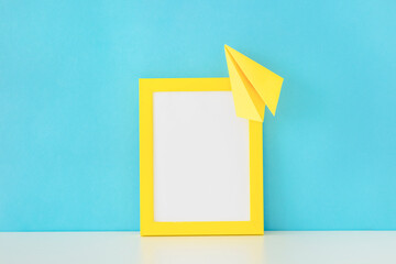 Bright yellow paper airplane soaring over a blank yellow picture frame against a blue background