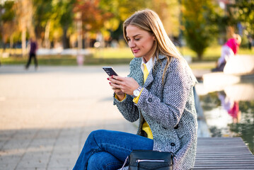 Portrait of modern businesswoman in stylish coat using smartphone while sitting on park bench in the city during sunny autumn day.	