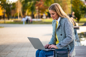Portrait of confident businesswoman in coat using laptop while sitting on a park bench in the city during sunny autumn day.	