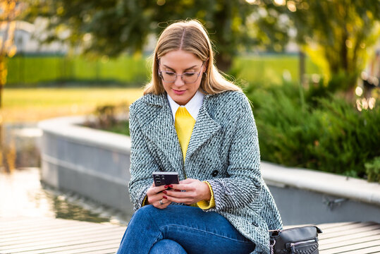 Portrait of urban businesswoman with eyeglasses in stylish coat using smartphone while sitting on park bench in the city during autumn day.	