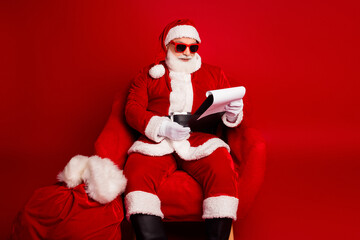 Santa in a bright red suit with sunglass sits in a plush chair reading papers against a solid red background creating festive holiday Christmas