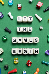 Wooden dice spell out 'Let The Games Begin' with a selection of colourful dice and game tokens lying around it on a vibrant green baize fabric