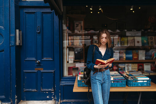 A young woman stands by a vibrant blue door outside a Camden, London bookstore, deeply engrossed in reading a red book. Books are displayed on a table.