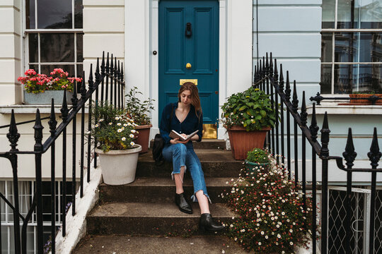 A young woman sits on the steps of a townhouse in London, reading a book surrounded by potted plants and flowers.