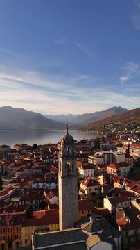 Cinematic drone flight over Lake Como framed by autumn-winter mountains and a red-tiled Italian village, with a tall historic bell tower rising above the rooftops