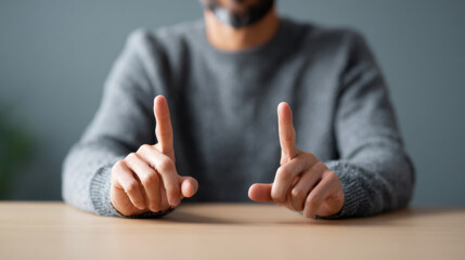 Close-up of man in gray sweater showing two index fingers pointing upwards on wooden table with blurred background