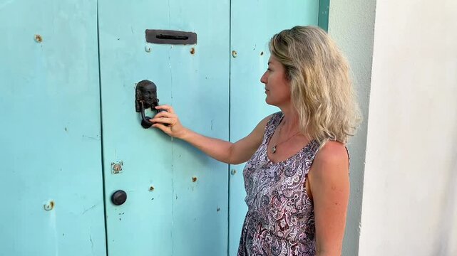  A side-profile, medium shot of a middle-aged woman with blonde, wavy hair wearing a patterned sundress. She is reaching out and holding a decorative, dark metal door knocker