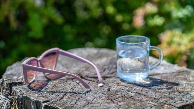 Pink sunglasses and glass of water on tree stump outdoors in summer sunlight