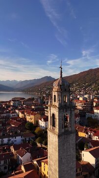 Aerial orbit of a historic stone bell tower rising above an Italian Lake Maggiore village, framed by red rooftops, autumn colors and alpine mountains in a serene cinematic atmosphere