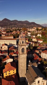 Aerial orbit of a historic stone bell tower rising above an Italian Lake Maggiore village, framed by red rooftops, autumn colors and alpine mountains in a serene cinematic atmosphere