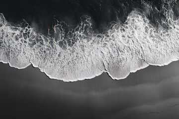 Aerial monochrome view of ocean waves crashing onto a dark sandy beach, creating intricate white foam patterns.