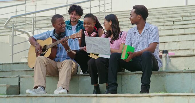 Indian young adult gen z lady woman male man group sitting on stair hold guitar play music sing song have fun joy. Happy pupil girl boy group spend break time relax mood clap hands enjoy new say place