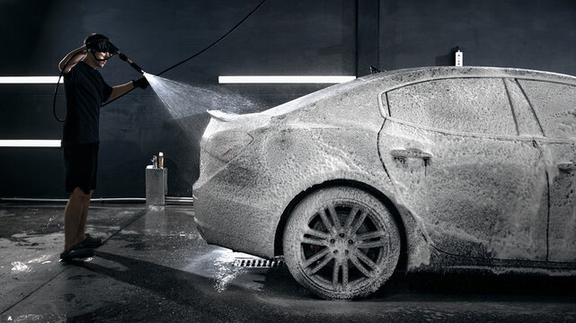 Worker spraying active foam on luxury sedan during car wash process. Concept of car wash detailing services, corporate communication, professional workflow quality.
