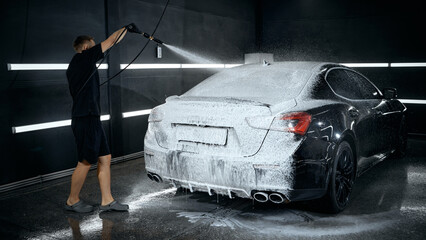 Worker applying active foam to black sedan under bright workshop light. Concept of car wash detailing services, franchise promotion, professional training visuals.