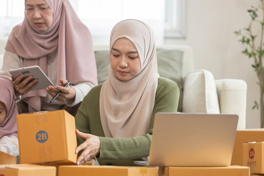 Muslim family running small online business from home, woman using laptop to manage orders surrounded by parcels. Concept of e commerce, home based startup and digital entrepreneurship.