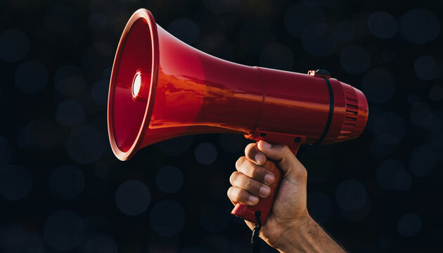 A person's hand firmly holds a bright red megaphone against a dark, blurred background, symbolizing the power of a single voice to make an important announcement