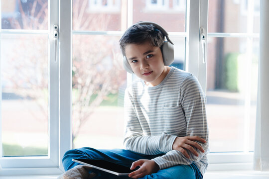 Smiling young boy wearing wireless headphones and holding a tablet while sitting by a bright window. Relaxed Preteen enjoying music or online learning at home in natural sunlight.