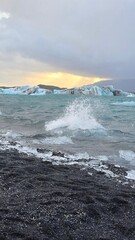 Waves crash onto the black sand shore of Jökulsárlón, scattering ice fragments as glowing icebergs float nearby under a dramatic Icelandic sky.