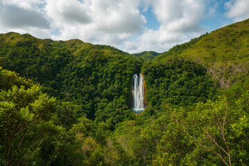 Scenic tropical waterfall surrounded by lush green rainforest, mountain hills under a partly cloudy sky. Perfect nature background for travel ads, eco-tourism brochures, posters, banners, website.