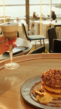 A hand holds a layered dessert coffee in warm sunset light beside a plate of stacked pancakes with nuts, captured in a cozy outdoor caf&eacute; setting.
