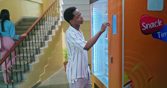 African young adult gen z male man select fast easy choose juice drink press button self service campus place. Happy pupil boy mobile phone use app look screen break time enjoy day indoor college area