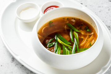 A close-up, high-angle studio shot of an Asian-style hot and sour noodle soup in a modern white bowl, served on an oval plate with condiments