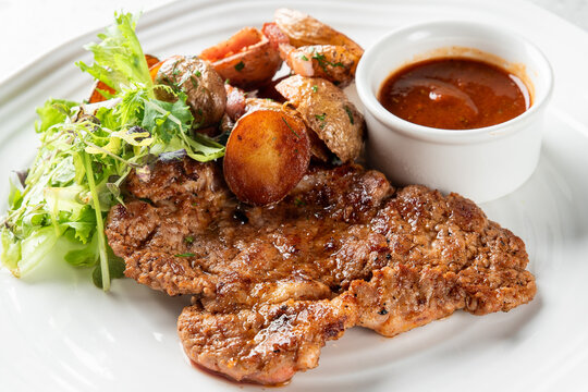 Appetizing pan-fried or grilled pork steak served on a white plate with roasted baby potatoes, a fresh green side salad, and a bowl of rich brown sauce