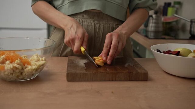 Close-up of a woman slicing a fresh peach on a wooden cutting board to prepare a healthy and delicious fruit salad in a modern kitchen, promoting a wholesome and nutritious lifestyle