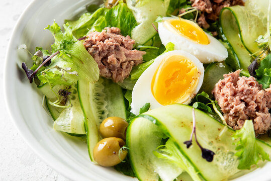 Close-up of a fresh, healthy tuna salad with cucumber ribbons, boiled eggs, olives, and mixed greens in a white bowl. Natural light, vibrant colors