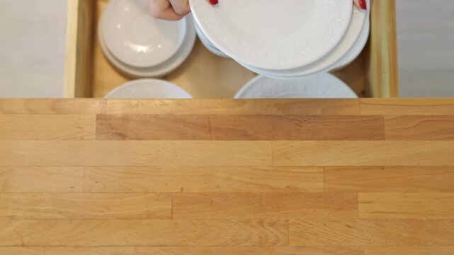 A woman with a red manicure is taking plates out of a drawer and placing them on a wooden surface