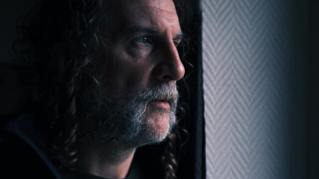 A contemplative man with curly hair and a mustache gazes out a window, bathed in soft natural light.