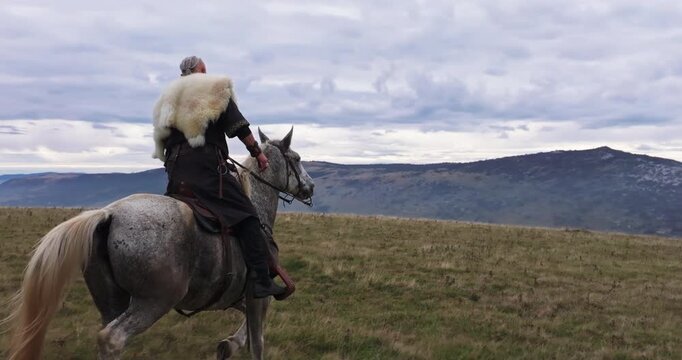 Aerial drone establishing or tracking shot of Bearded warrior in fur cloak riding a white horse across open landscape at sunrise, mountains in the background. Historical, rugged, adventurous scene
