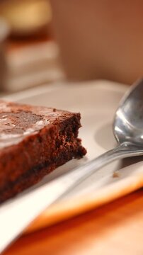 A piece of chocolate cake is on a white plate with a spoon next to it. The cake is cut into a triangle and has a frosting on top. The spoon is silver and is placed on the right side of the plate