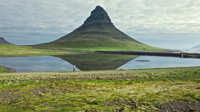 Tourist walking by Kirkjufell mountain with reflection in calm water, Sn&aelig;fellsnes Peninsula, Iceland. Iconic landscape and travel destination captured in scenic summer light.