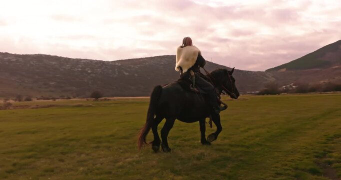 Cinematic aerial drone shot of a Viking warrior on a large black horse surveys a dramatic landscape during sunset, wearing a fur cloak concept of Ancient exploration, adventure, and the spirit of the